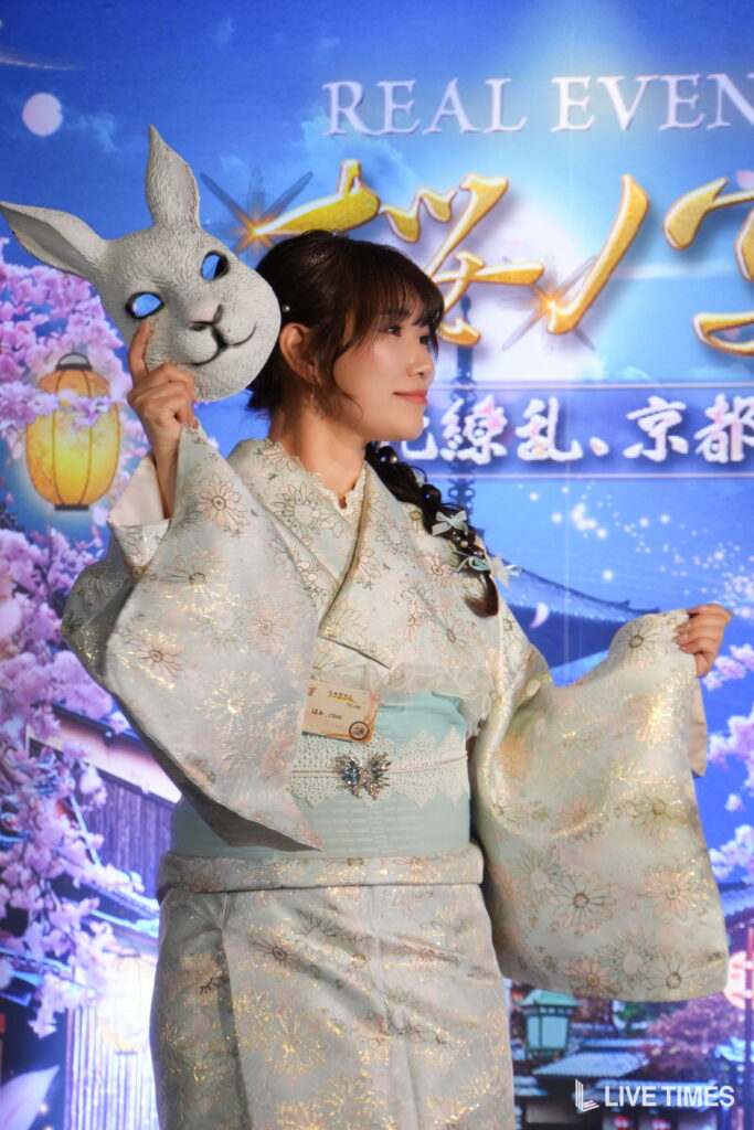 Young woman in a pale blue floral kimono holding a gray rabbit mask beside her face at a colorful festival backdrop.