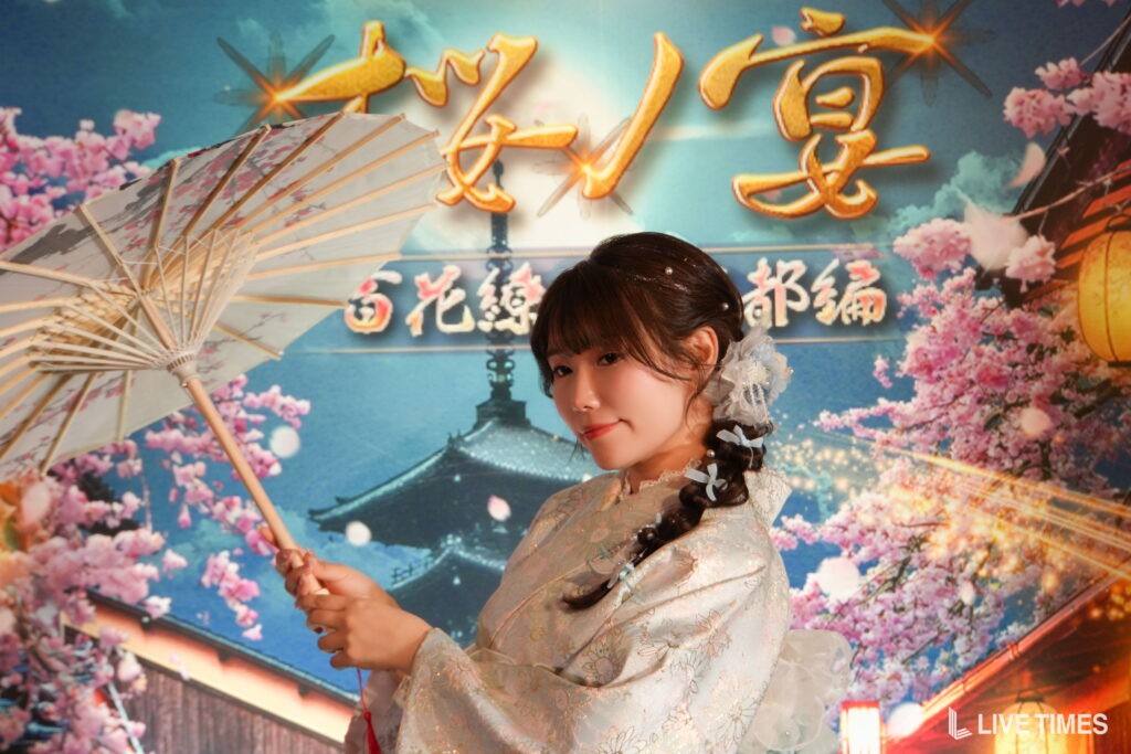 Portrait of a young woman in a pale floral cheongsam holding a paper fan, with a cherry blossom backdrop behind her.