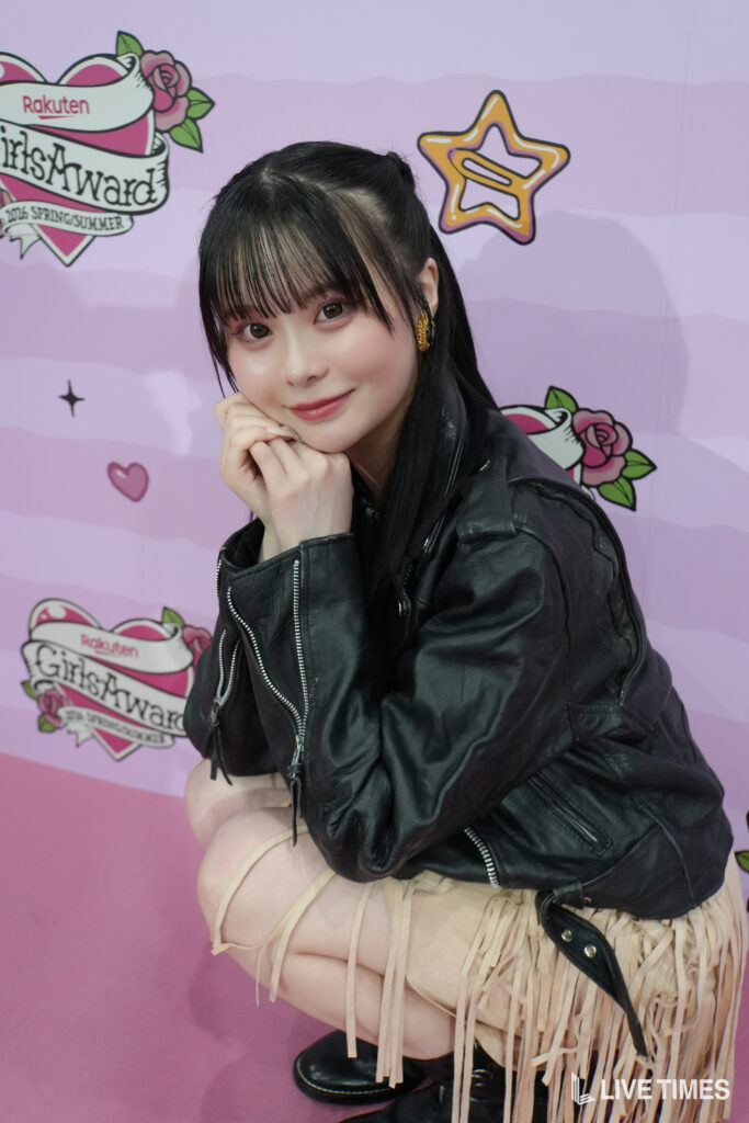 Young woman with black hair in a ponytail, wearing a black leather jacket, posing with hands under chin against a pink backdrop with event logos (GirlsAward).