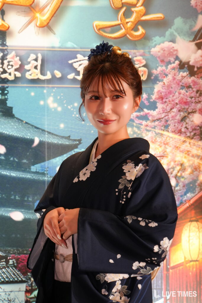 Portrait of a woman in a navy kimono with white floral embroidery, standing before a backdrop of cherry blossoms and lights.