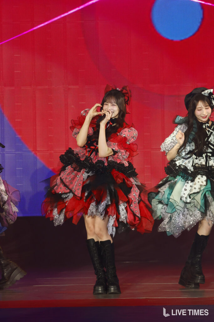 Two performers in frilly, colorful stage outfits sing into microphones on a bright red stage backdrop; one in red and black, the other in teal and silver dresses, both wearing knee-high boots.