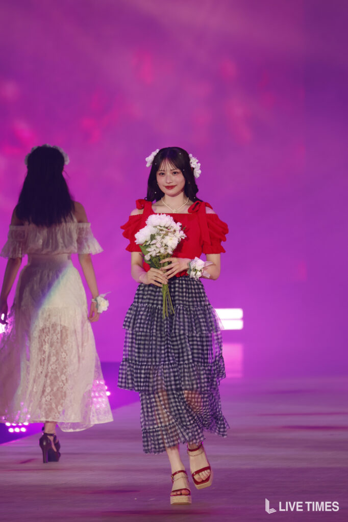 Front-facing woman on a stage wearing a red off-shoulder top with ruffles and a layered navy gingham skirt, holding a bouquet of white flowers, with floral headpiece under pink lighting.