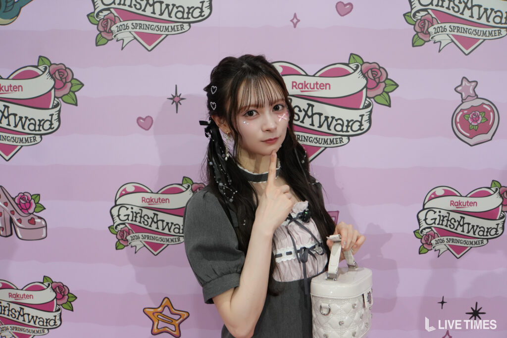 Young woman posing at a pink GirlsAward backdrop, wearing a gray dress and holding a white handbag, with finger to lips gesture.