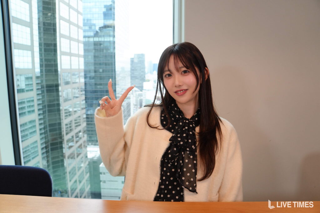 Woman in a cream coat with a black polka-dot scarf, smiling and making a peace sign at a desk near a large window overlooking city skyscrapers in an office setting, Live Times watermark visible in corner.