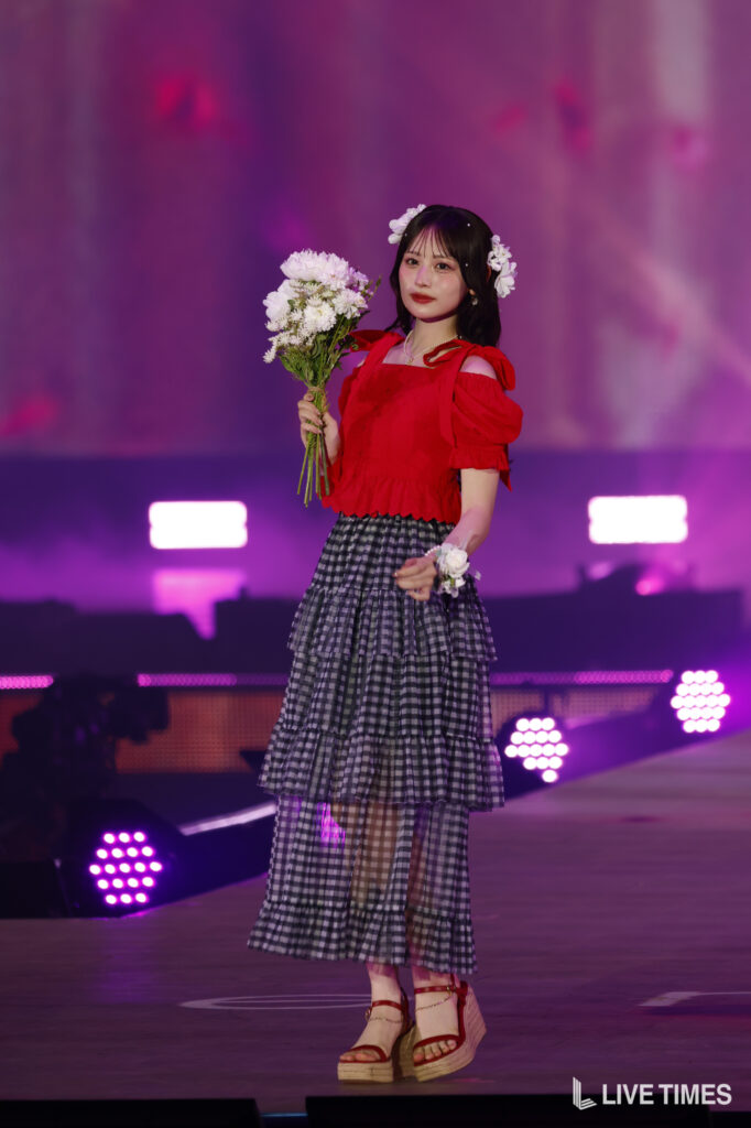 Performer in a red off-shoulder top and tiered plaid skirt holding a bouquet on a colorfully lit stage.