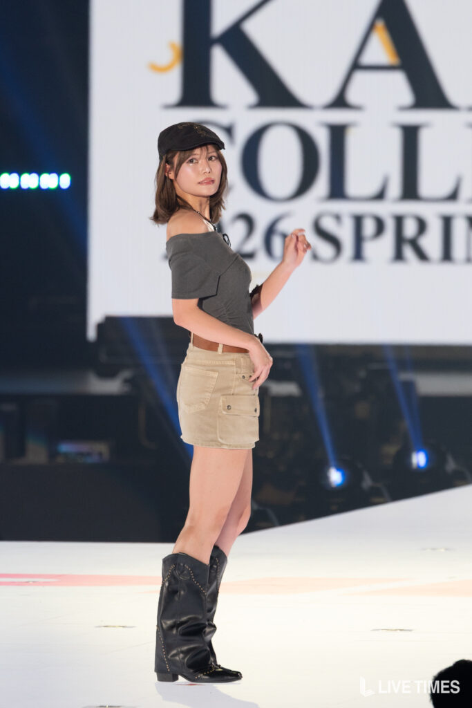 Female model on a stage posing in a gray off-shoulder top, beige cargo skirt, black hat and tall black boots.