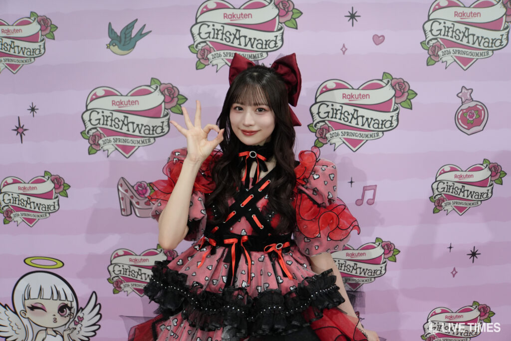 Person in a red and black frilly dress making an OK gesture in front of a pink GirlsAward backdrop with logos and hearts.