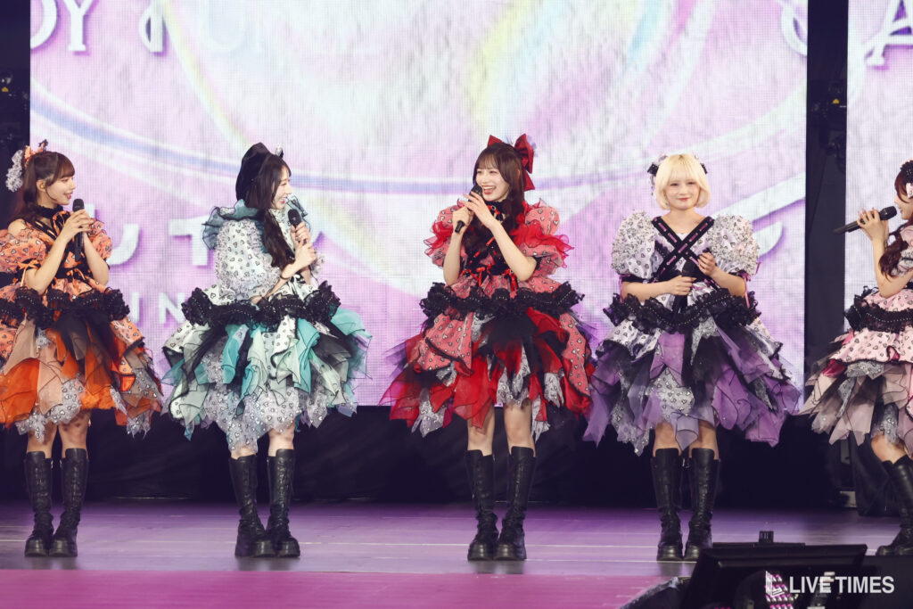 Five performers in colorful, layered ruffled dresses and knee-high boots sing into microphones on a bright stage backdrop.