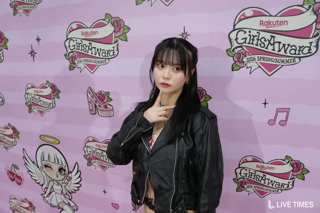 Woman in a black leather jacket posing with finger on chin in front of a pink GirlsAward backdrop.
