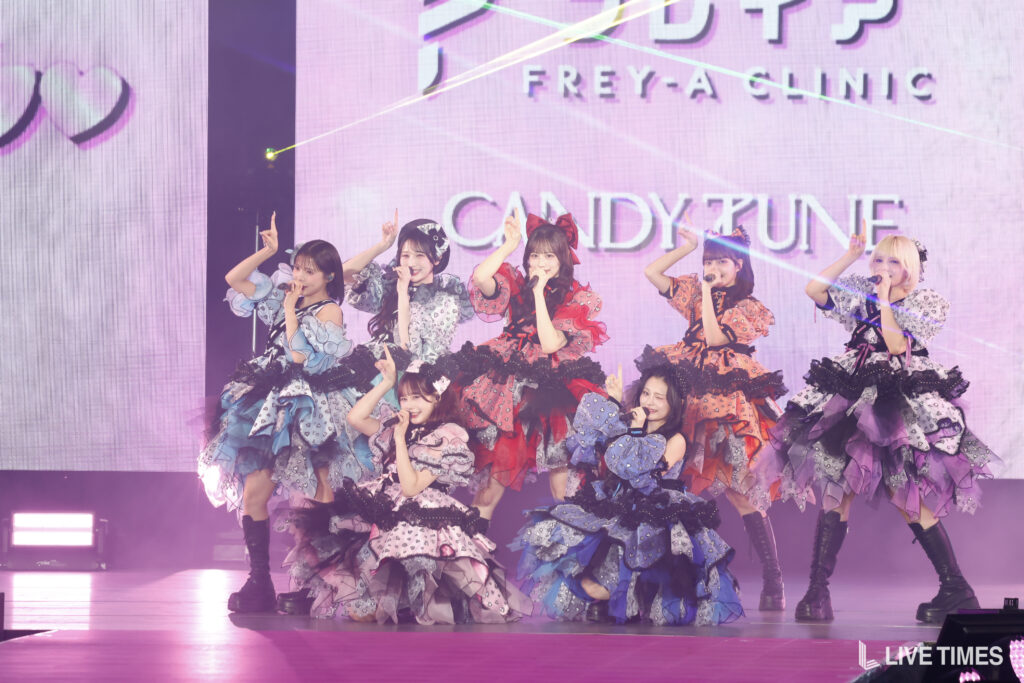 Eight female idols in colorful, ruffled outfits perform a choreographed stage routine with microphones under purple lighting.
