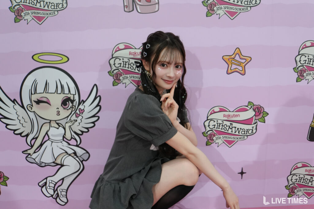Young woman poses with finger to her cheek in front of a pink GirlsAward backdrop with cartoon angels and logos.