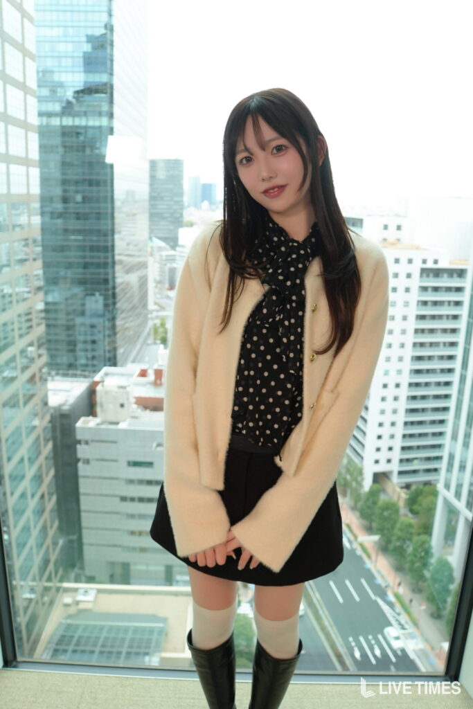 Portrait of a young woman with long dark hair, wearing a cream coat and black polka-dot blouse, standing by a glass window with a city skyline behind her.