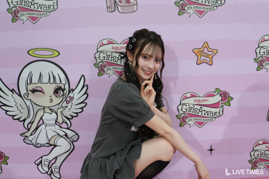 Young woman posing with hand near her face in front of a pink backdrop featuring cartoon angels and 'GirlsAward' logos (2016 Spring/Summer).