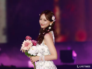 Woman in a white floral dress holding a pink bouquet on a dimly lit stage with purple lighting.