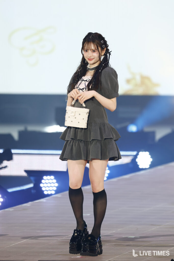 Woman in a gray tiered dress on stage, holding a small white handbag, with black knee-high socks and platform shoes.