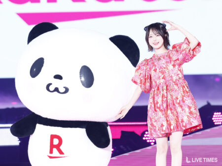 Woman in a pink floral dress posing with a giant panda mascot on stage at a live event.