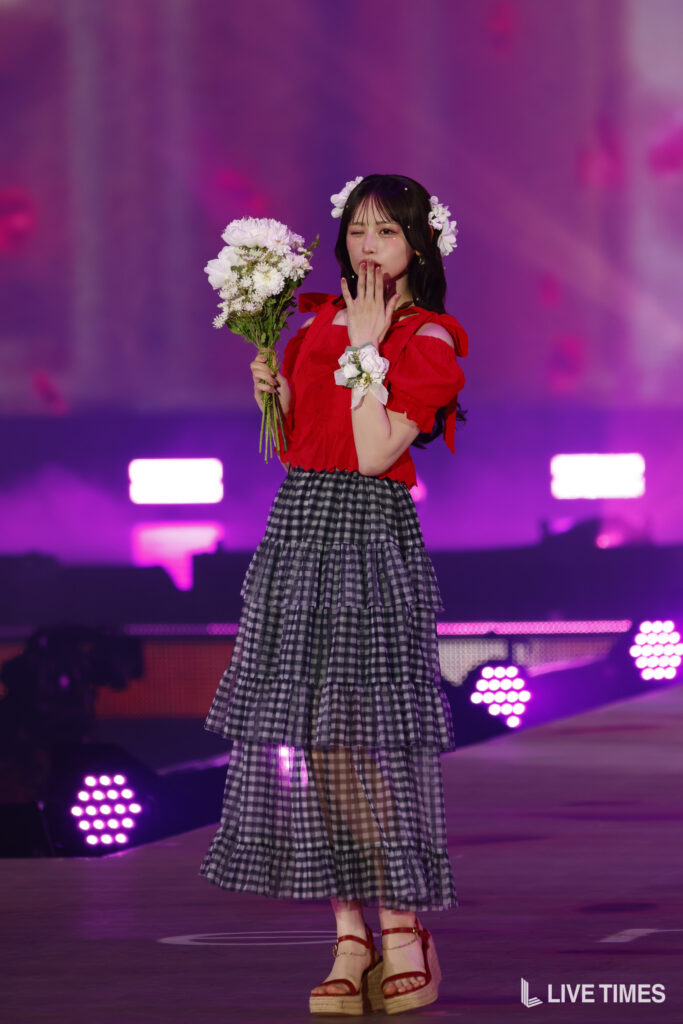 Performer on a purple-lit stage wearing a red blouse and tiered black-and-white gingham skirt, holding a bouquet and blowing a kiss.