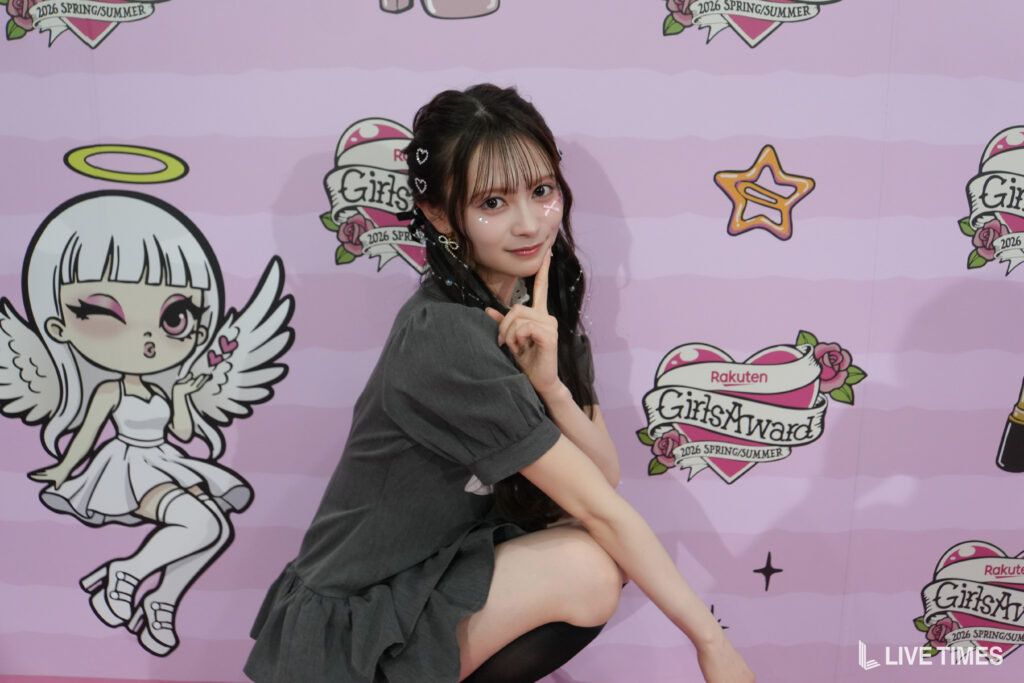 Young woman in a gray dress poses with a finger to her lips against a pink backdrop featuring cartoon angels and 'GirlsAward' logos.