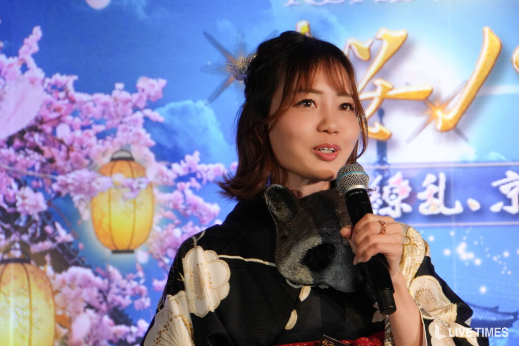 Asian woman in a kimono speaking into a microphone at a stage with a blue backdrop and pink cherry blossoms behind her.