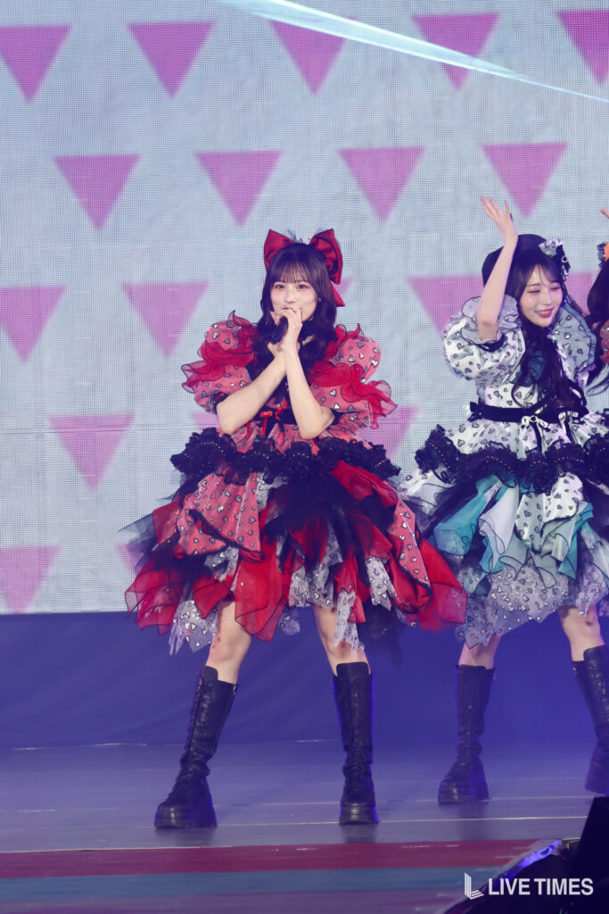 Two female pop idols performing on stage in elaborate frilly dresses; center singer in red and black with a large red bow sings into a microphone, a second dancer in gray-blue raises her arms beside her; both wear knee-high boots.