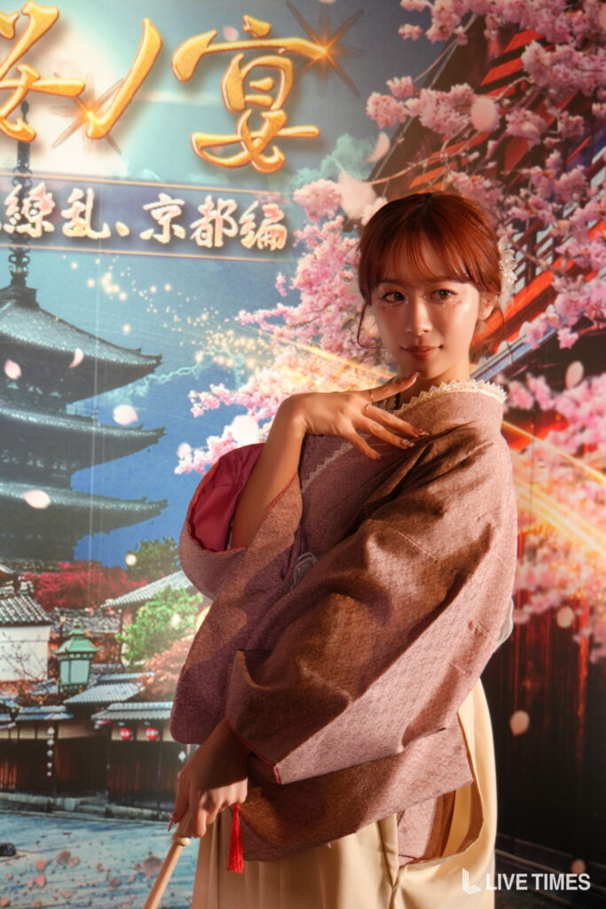 Young woman in a pink kimono posing with her hand under her chin, in front of a cherry blossom backdrop with a pagoda/temple scene.