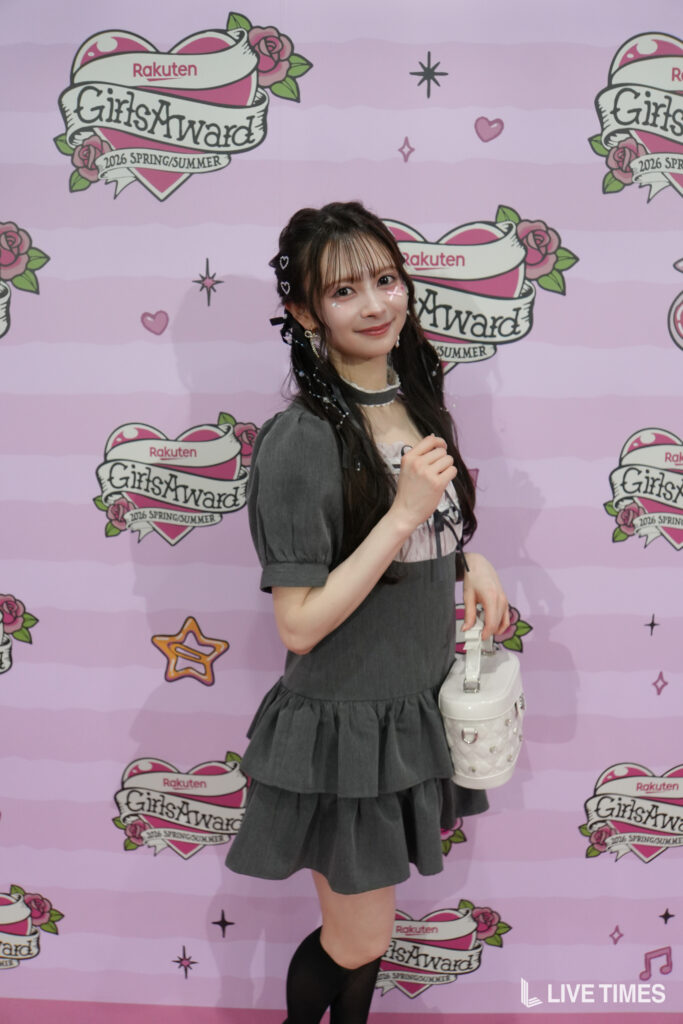 Young woman posing at a pink backdrop with Rakuten logos and GirlsAward branding, wearing a gray dress and holding a white handbag.