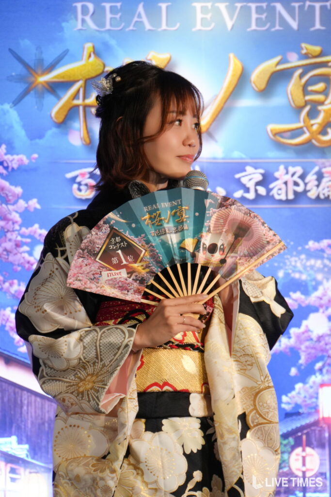 Woman in a patterned kimono with a decorative hair accessory, holding a colorful folding fan amid a blue event backdrop and microphone in hand.