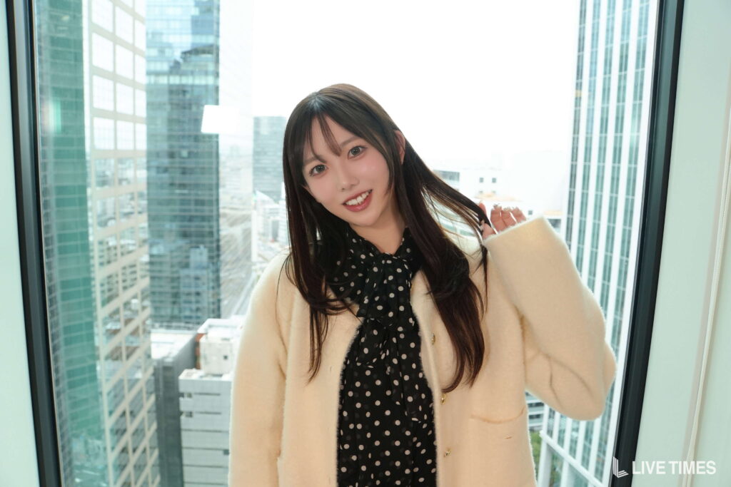 Young woman with long dark hair smiling at the camera, wearing a black polka-dot scarf and cream coat, in front of a large window with a city skyline outside.