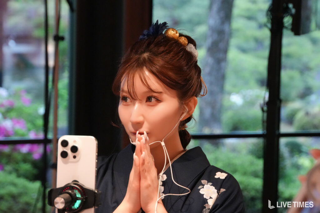 Woman in a dark floral kimono looks at her phone while wearing white wired earphones, hands clasped near her face, with a microphone in front.