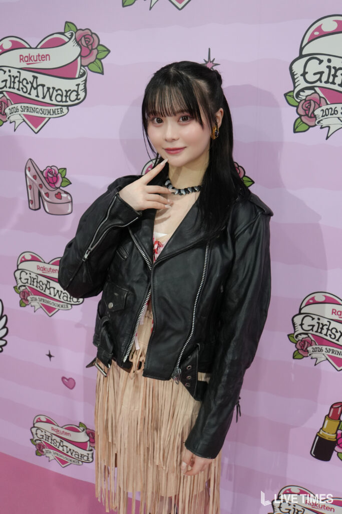 Young woman with black hair in a black leather jacket and beige fringe dress posing in front of a pink GirlsAward backdrop.