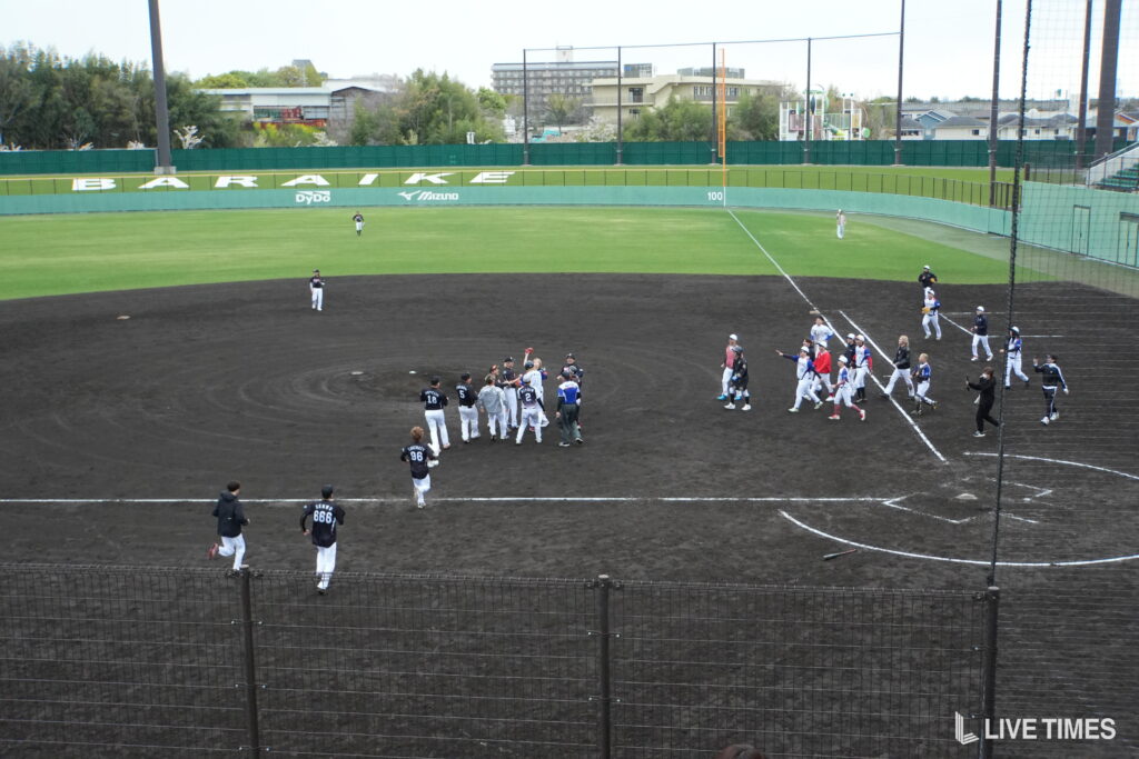 Baseball players in dark and white uniforms huddle on the infield dirt as teammates walk in from the right field line. Behind them is the green outfield wall and stadium seating.