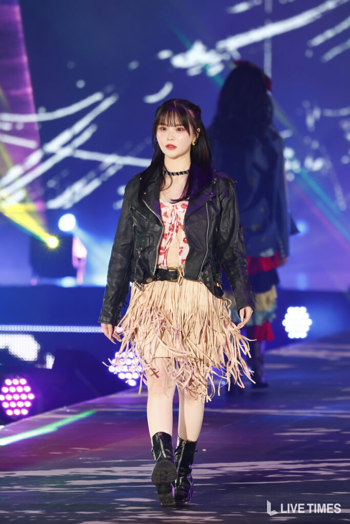 Young woman walking a stage in a black leather jacket and fringed beige dress, wearing boots, under colorful stage lights.