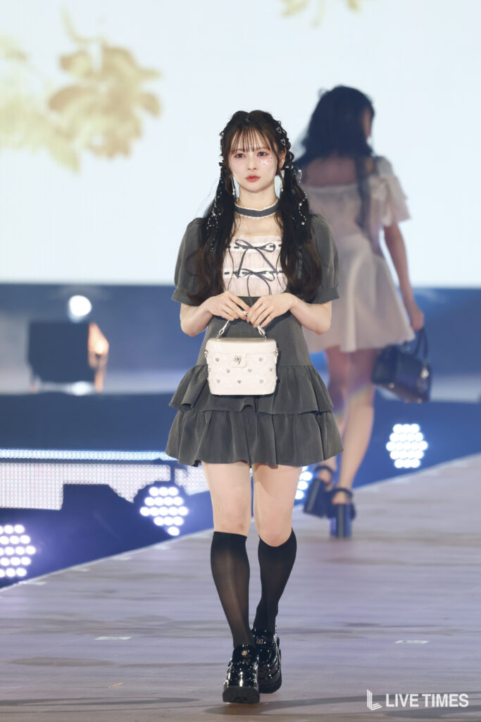 Runway model walking in a gray layered dress, black knee-high socks, shiny black shoes, and a small white studded handbag.