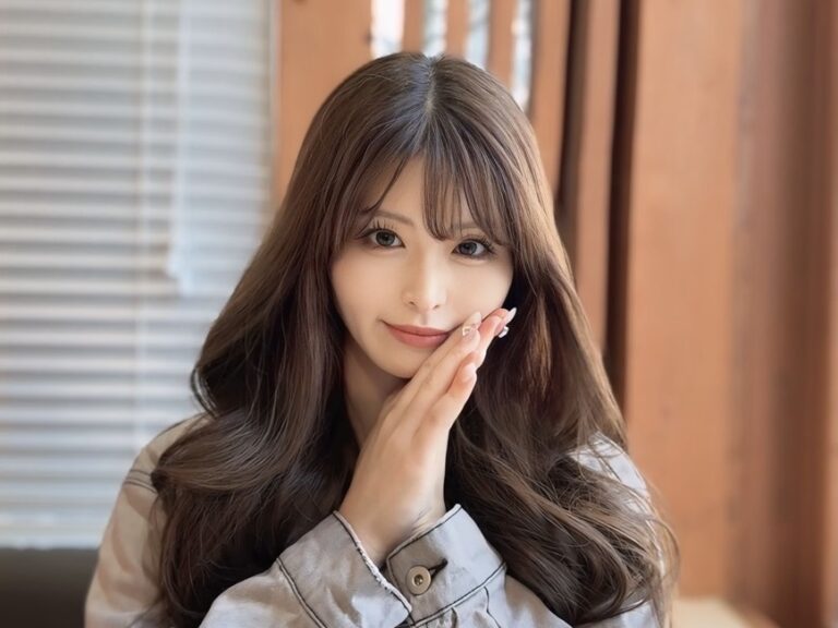 Young woman with long wavy hair and soft smile, hands pressed together near her face in a warm, indoor setting.