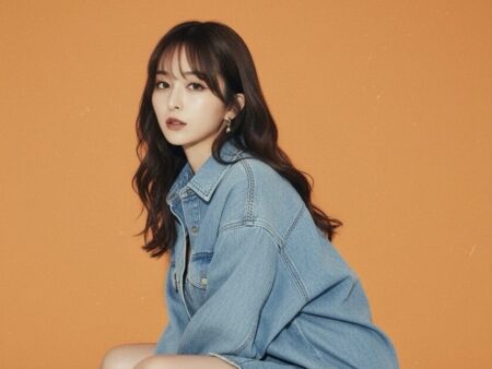 Woman wearing a light blue denim jacket, seated against an orange backdrop, looking at the camera.