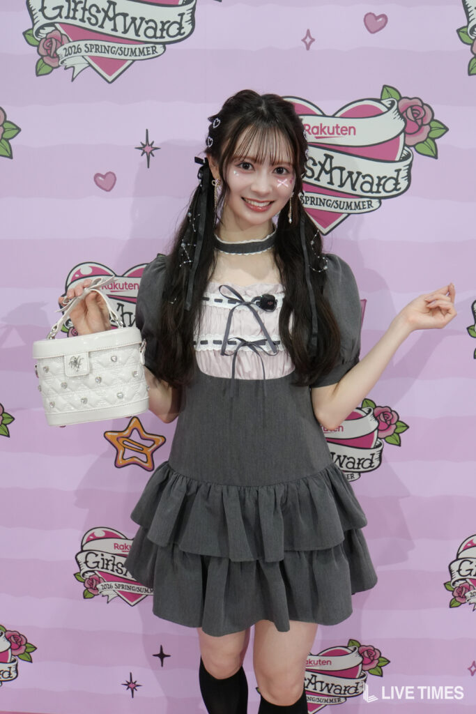 Young woman in a gray dress holding a small white quilted handbag in front of a pink backdrop with 'GirlsAward' logos, smiling at the camera.