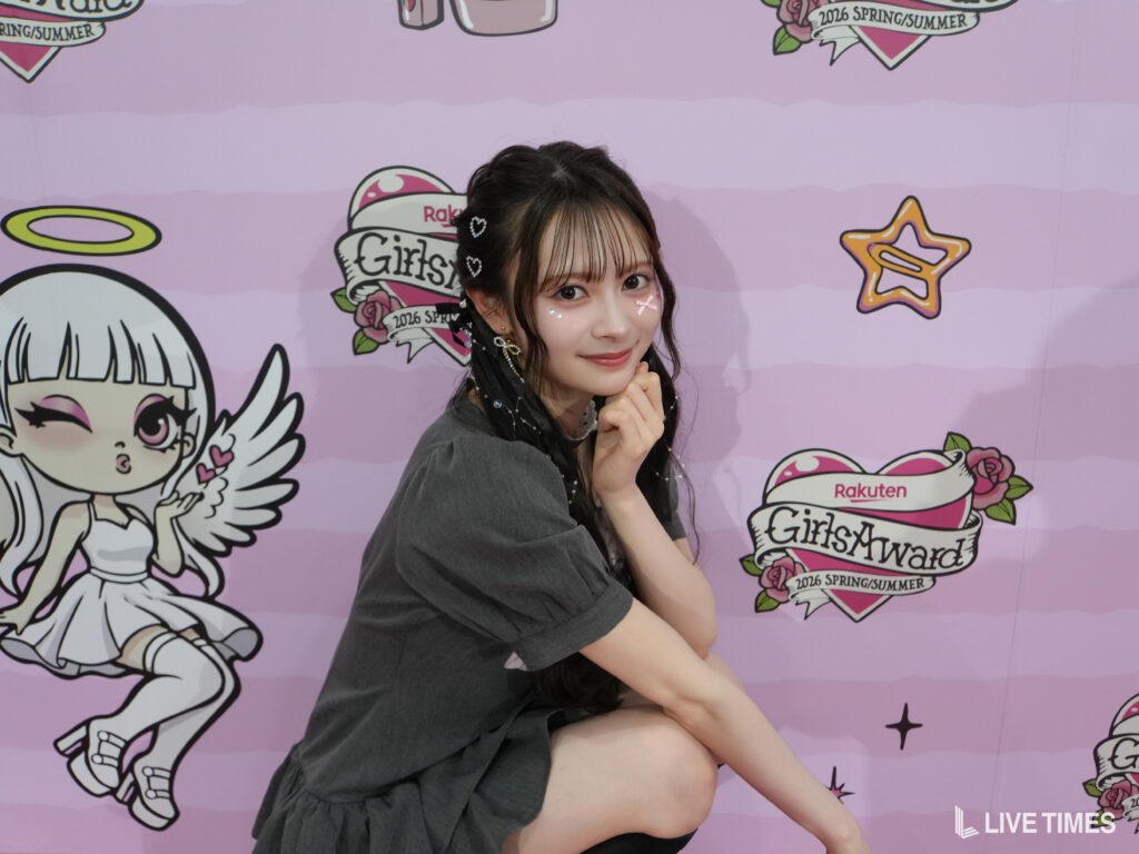 Young woman posing against a pink event backdrop with cartoon graphics and 'GirlsAward' logos, touching her chin with a gentle smile.