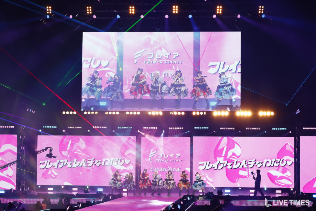 Girls' idol group performing on a large stage with bright pink screens and multicolored spotlights.