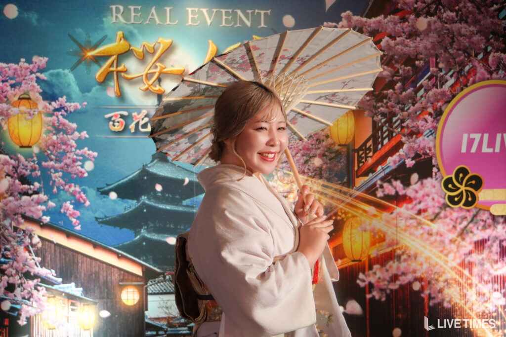 Smiling woman in a light outfit holding a floral parasol with a cherry blossom background behind her.