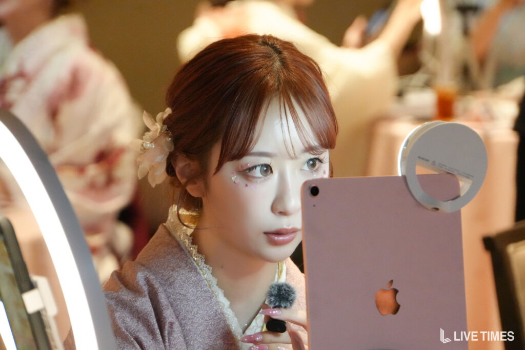 Life-like mannequin in makeup posing for a mirror selfie at a vanity with a pink smartphone and ring light behind.