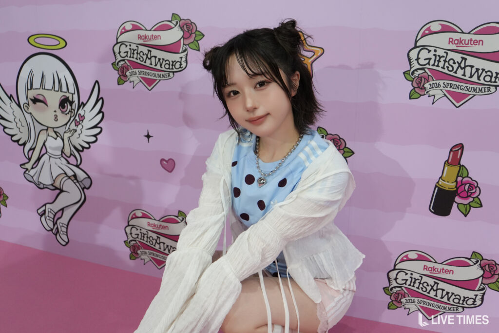 Young woman posing in front of a pink backdrop with GirlsAward logos, wearing a blue polka-dot top and white cardigan.
