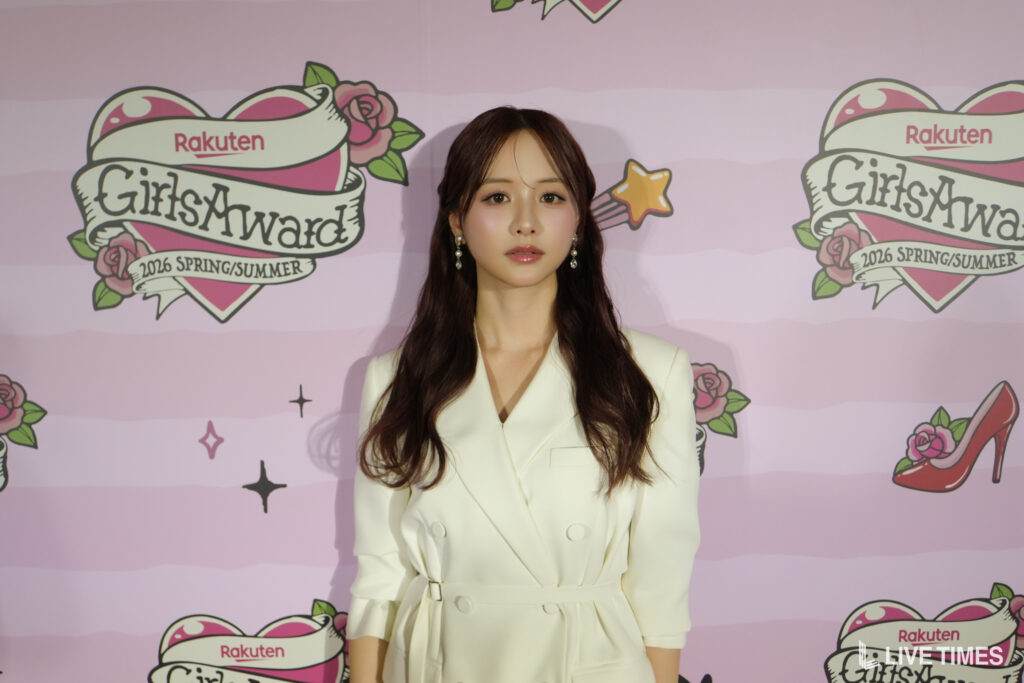 Young woman with long dark hair wearing a white blazer posing in front of a pink backdrop featuring the Rakuten GirlsAward logo and decorations.