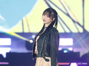 Female performer on stage wearing a black leather jacket and light skirt, looking toward the audience with colorful stage lights behind her.