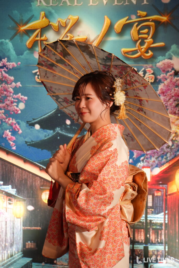 Woman in a pink floral kimono holding a decorative parasol, posing in front of a colorful festival backdrop with blossoms and Chinese lanterns.