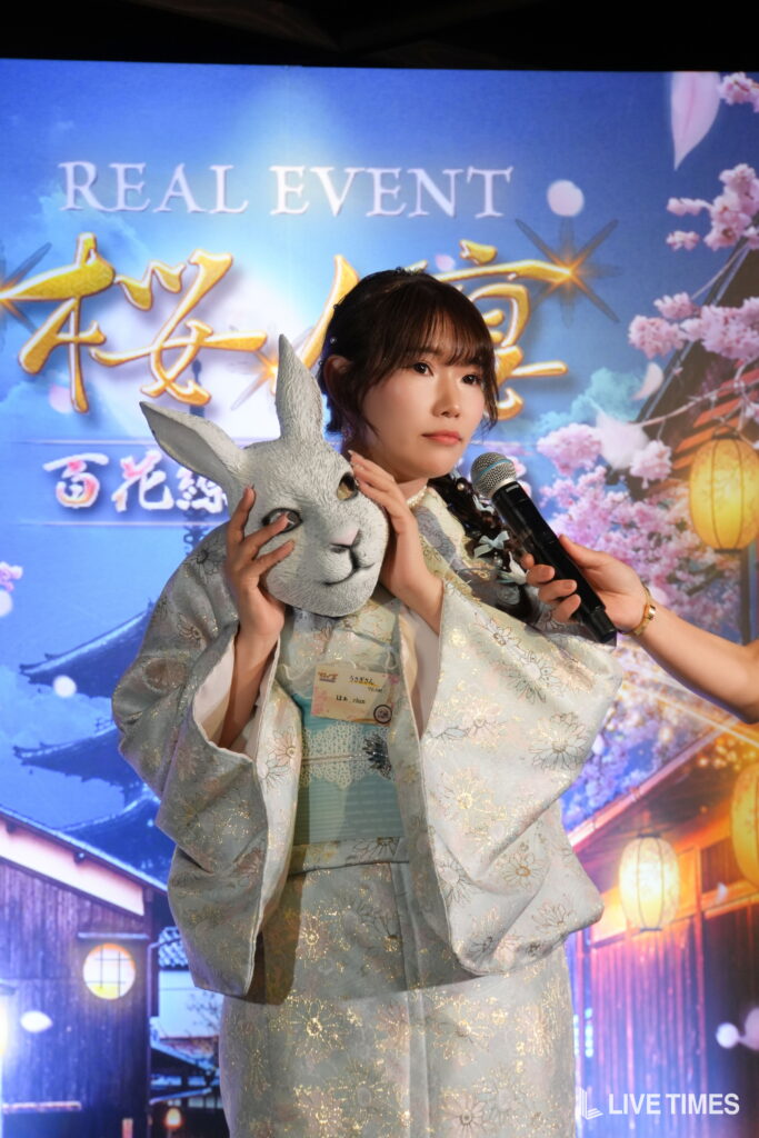 A woman in a light blue patterned kimono holds a gray plush rabbit mask while a hand with a microphone interviews her on stage.