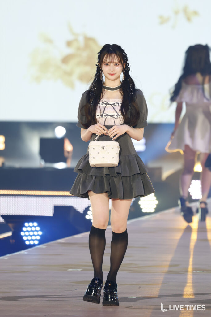 Runway model in a gray tiered dress with black knee-high socks, patent shoes, and a white studded handbag.