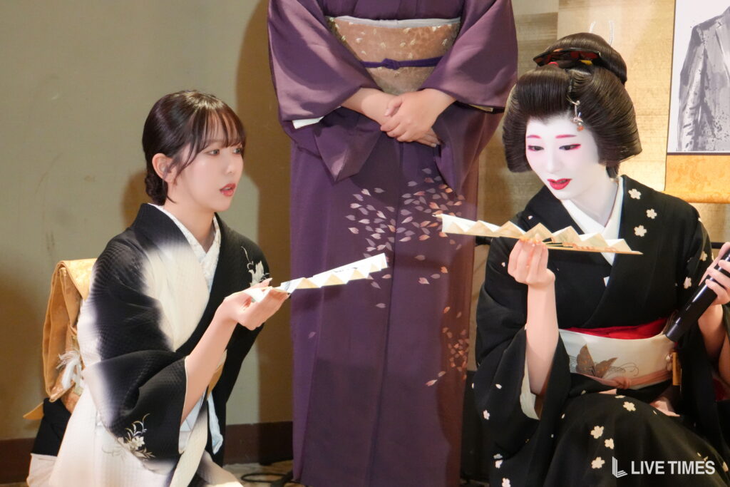 Two women in traditional kimonos with white makeup hold folded paper fans while a person in a purple kimono stands behind them, suggesting a traditional performance scene.