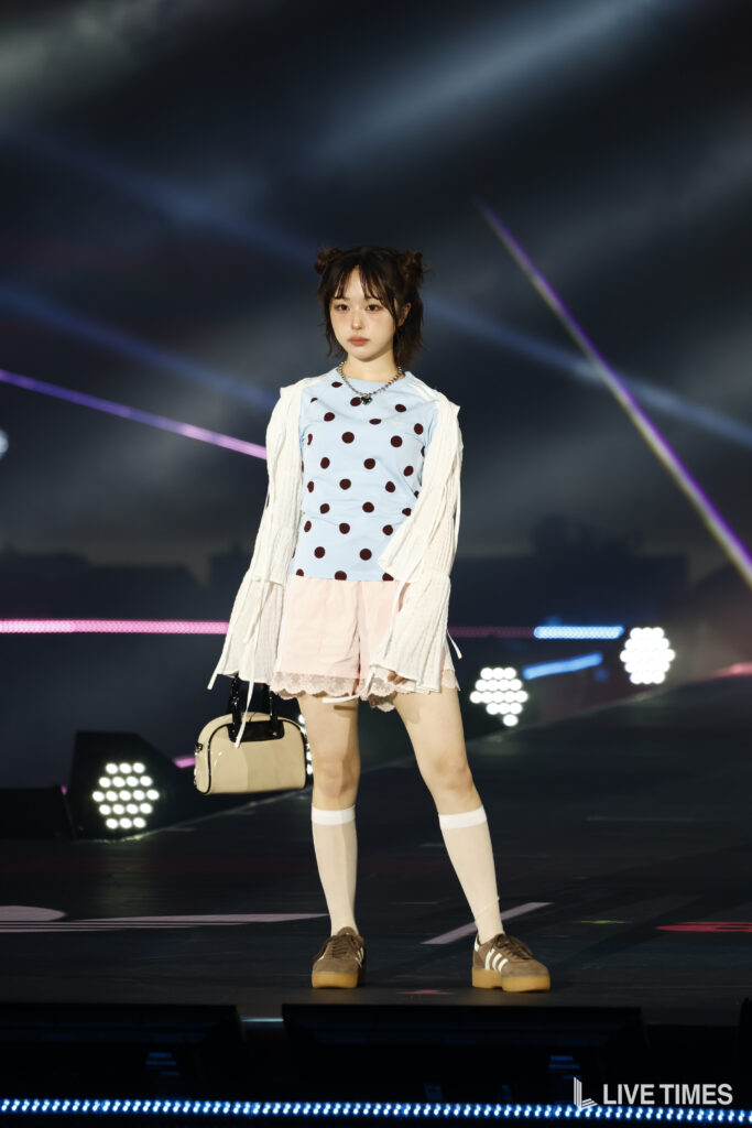 Stage performer standing on bright-lit runway in a pale blue polka-dot top, cream cardigan, pink skirt, knee-high socks, and a beige handbag.