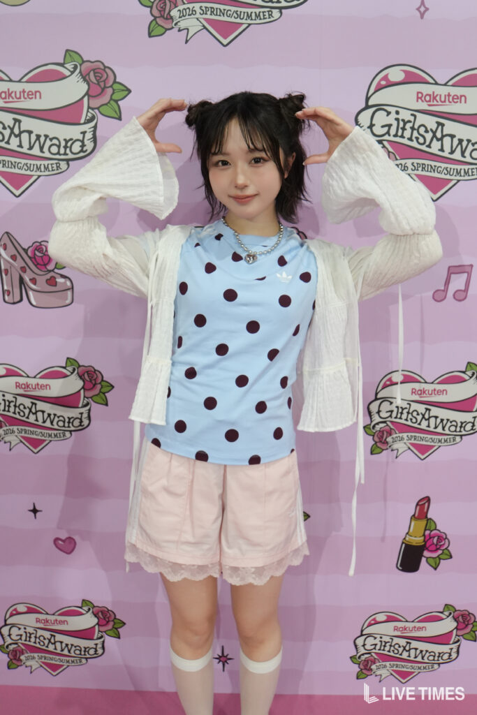Young woman with short dark hair in pigtails making a heart shape with her hands, wearing a light blue polka-dot top and pale pink shorts in front of a pink event backdrop.