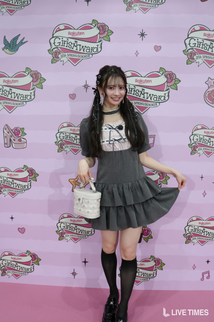 Young woman in a gray, layered dress posing with a white handbag in front of a pink GirlsAward backdrop.